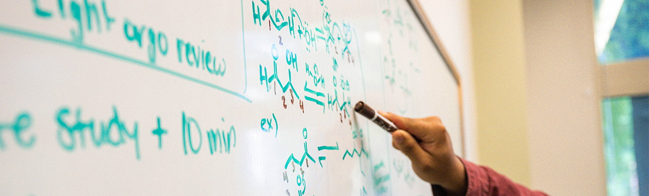 Close-up on a hand writing scientific equations on a whiteboard.