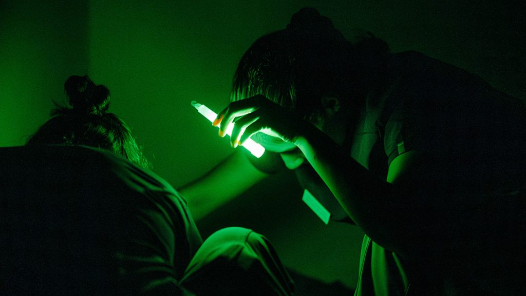 A UNCG nursing student uses a glowstick to check a patient in a dark room.