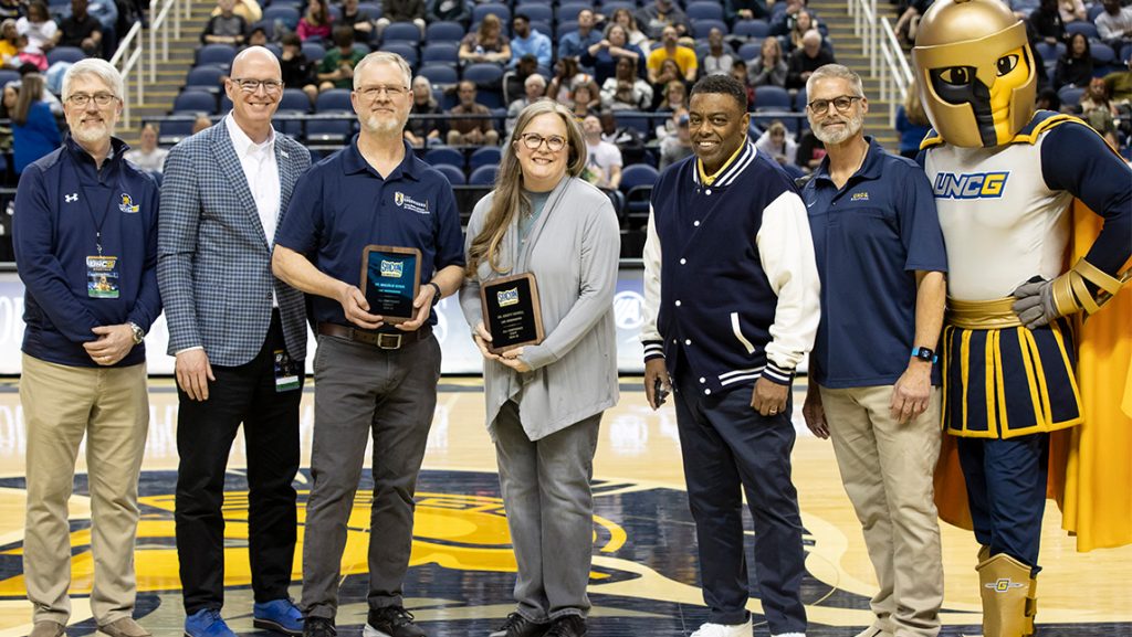 Malcolm Schug and Kristy Howell receive awards with UNCG Chancellor Gilliam, Provost Boyette, and Spiro beside them on the basketball court.