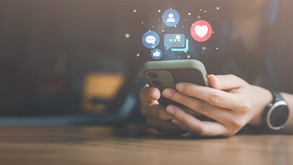 Close up on hands holding a phone with social media reaction emojis bursting out.