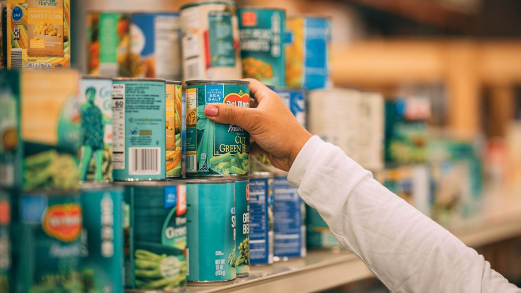 A hand reaches for canned food off a shelf.