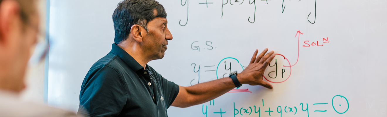 A professor stands in front of the white board pointing at and explaining an equation