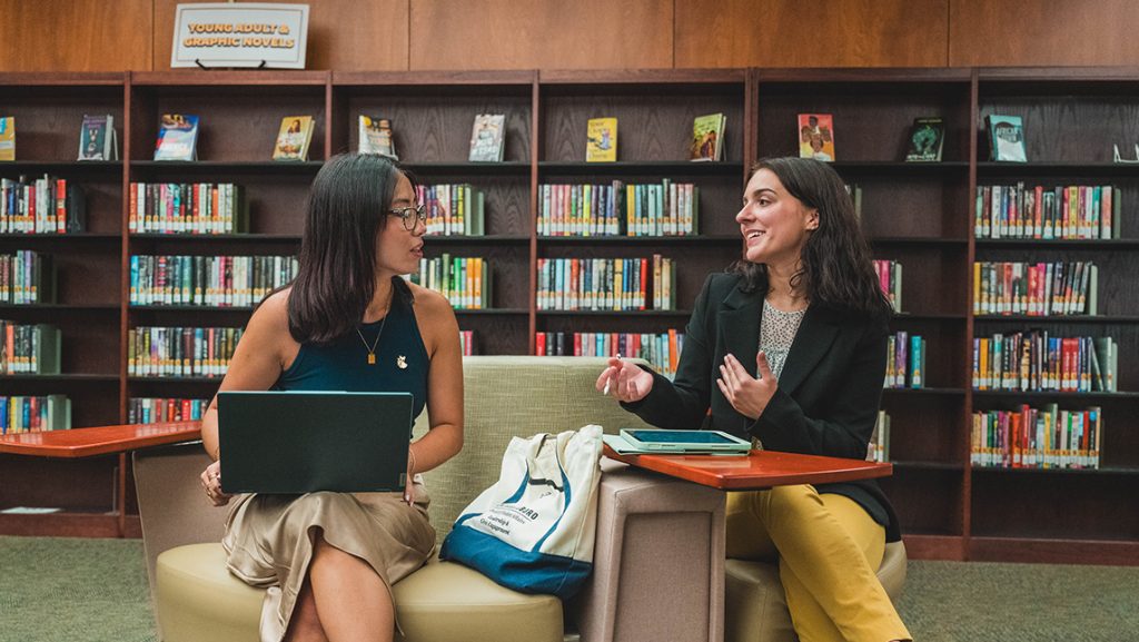 Two students have a conversation in a library with laptops and tablets. 