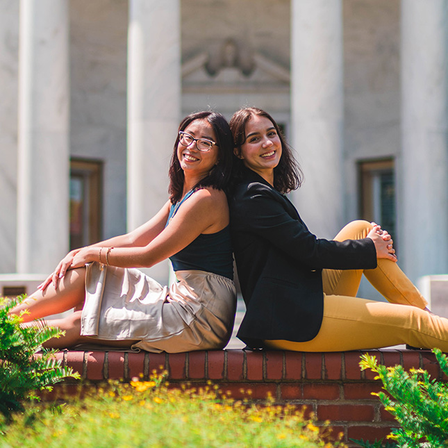 Two female students sit back to back on a brick ledge in front of Jackson Library.