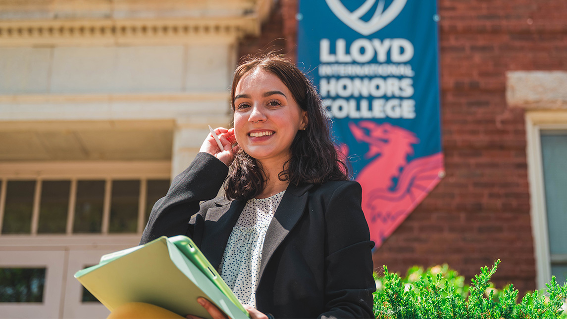 Student with laptop stands in front of a campus building with a Lloyd International Honors College banner hanging on it. Student
