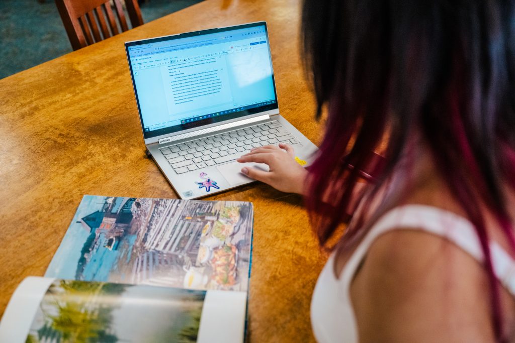 A student sits at a wooden table with a laptop and open textbook in front of them.