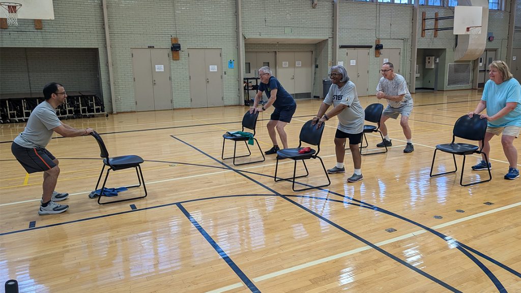 UNCG kinesiology student leads senior citizens holding onto chairs in exercise.