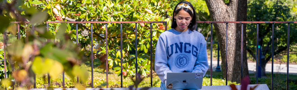 A student wearing a UNCG sweatshirt sets outside working on their laptop. Fall leaves are at the forefront of the image.