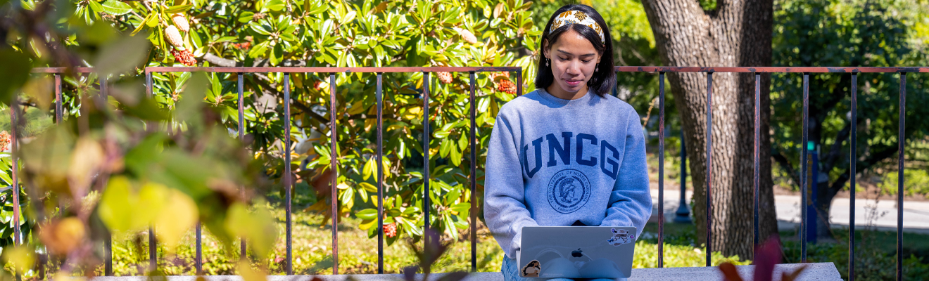 A student wearing a UNCG sweatshirt sets outside working on their laptop. Fall leaves are at the forefront of the image.