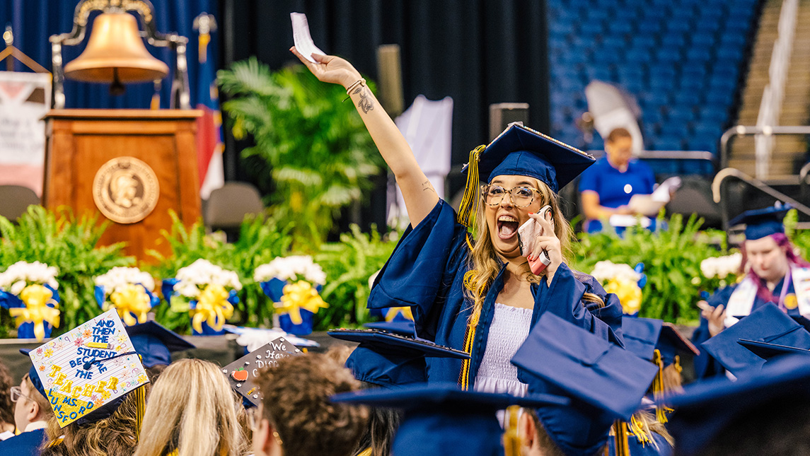 Graduate holds her arm up and screams happily in the phone as she finds her parents in the audience.
