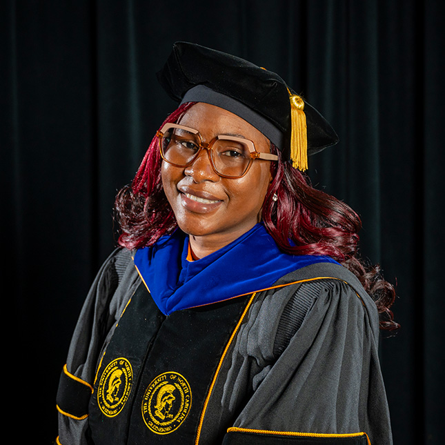 Doctoral grad poses in cap and gown in a black studio.