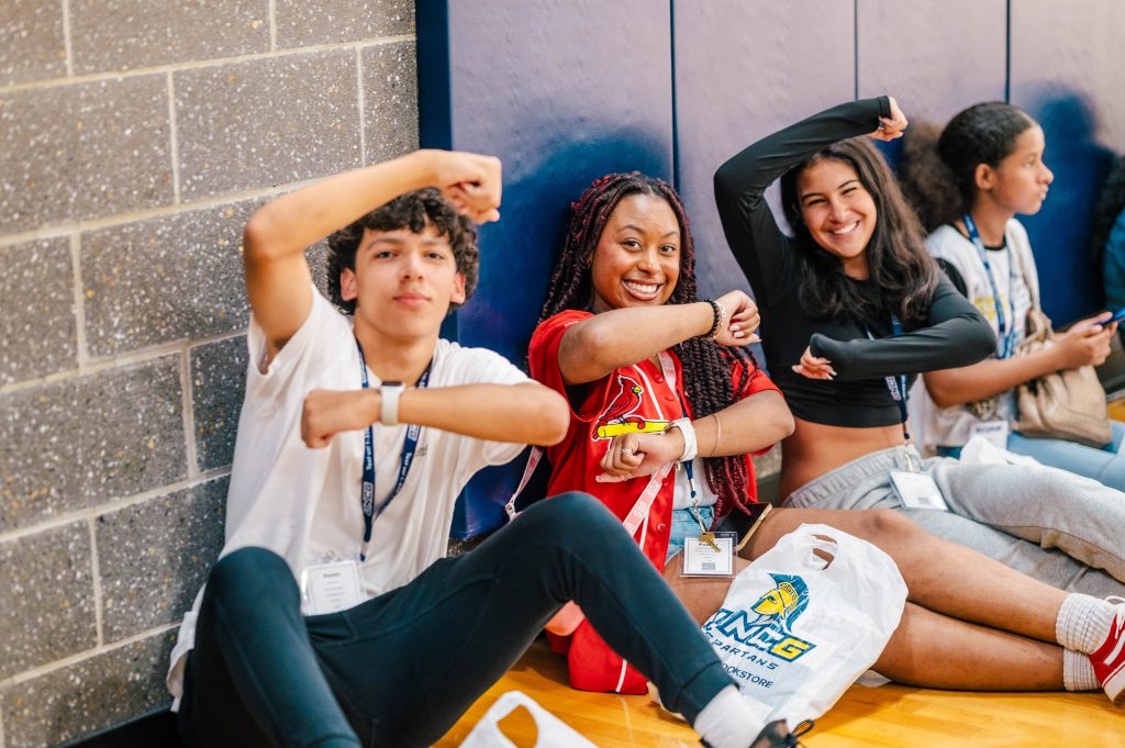 Three students sit on a gym floor and make a G with their arms.