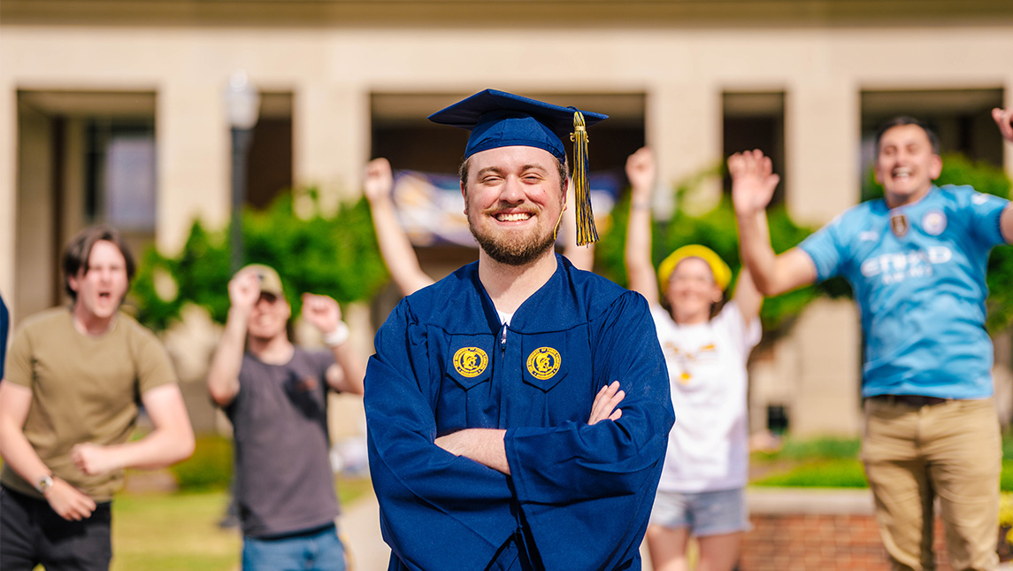 Student in cap and gown stands in front of cheering friends in front of the EUC.