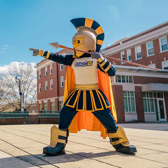 A Spartan mascot stands in front of a campus dorm and points towards the quad with a spear.