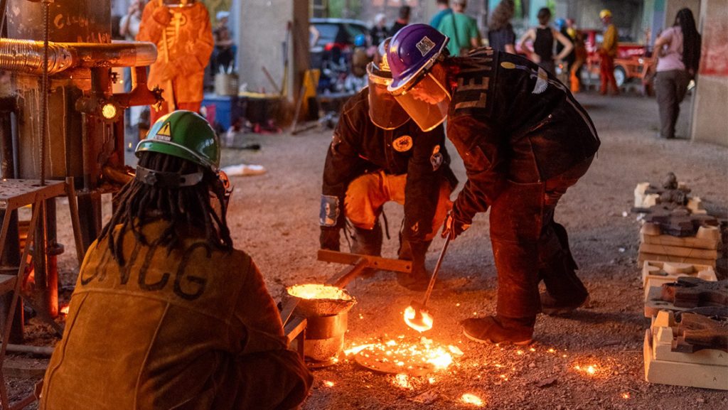 UNCG student bends over iron work still glowing from the fire.