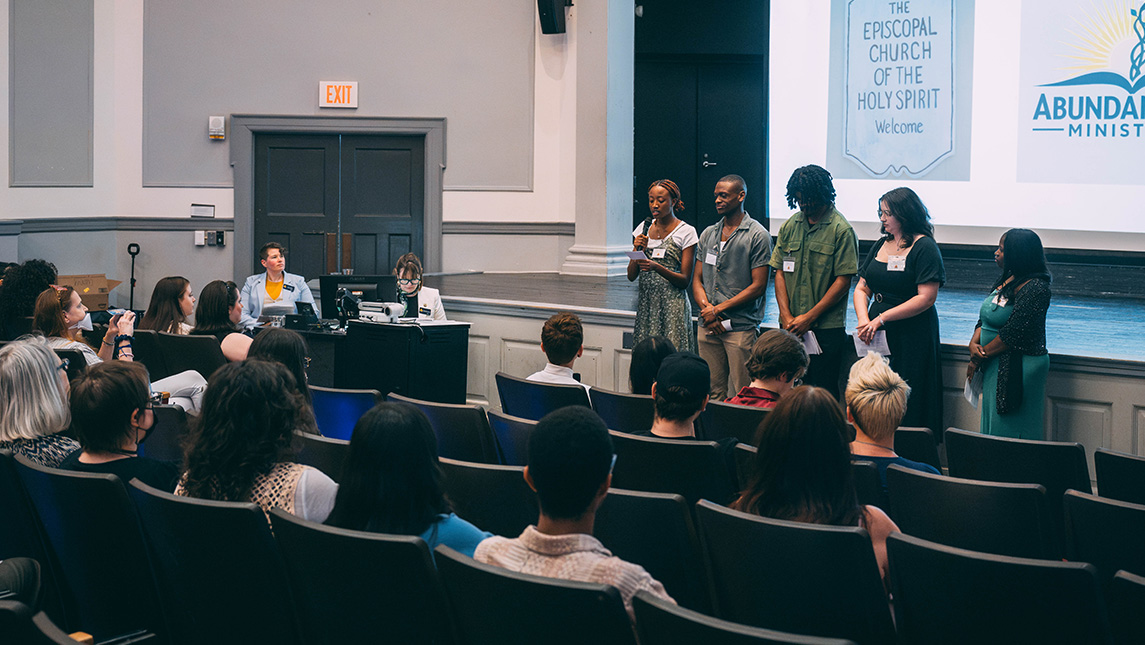 Group of students stand in front of folks seated in an auditorium with a screen behind them showing logos from Abundant Life Ministries.