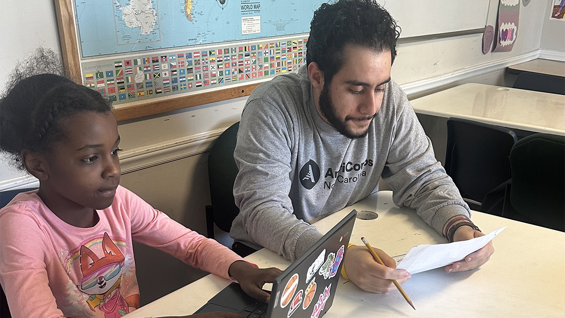 A Center for New North Carolinians volunteer works with a young student at a laptop.