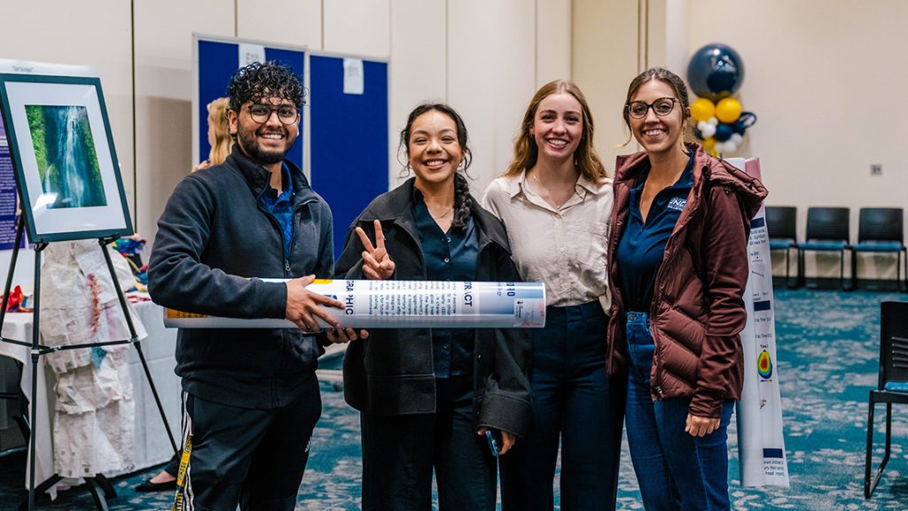 Four UNCG students hold up a rolled-up poster.