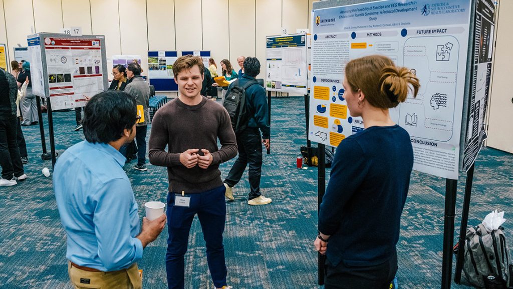 Three UNCG students talk next to a poster on display.