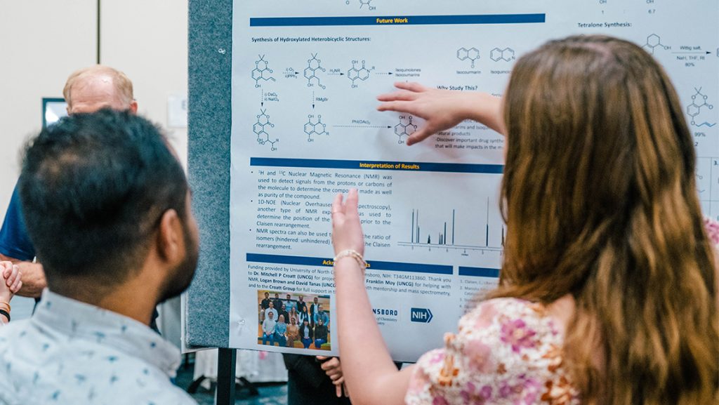 A UNCG student points to a graph on a research poster at a presentation.