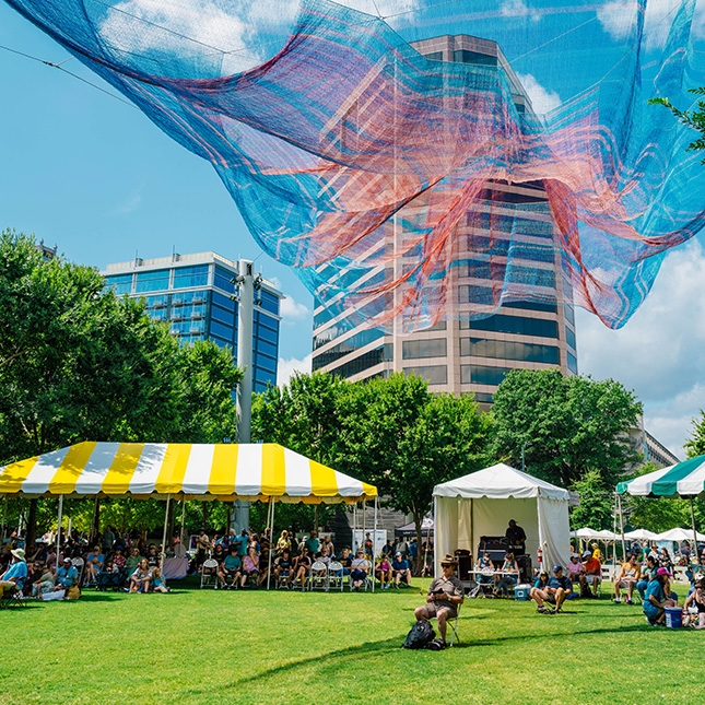 LeBauer Park lawn with people sitting under tents and a colorful gauze artwork hanging above with buildings behind.
