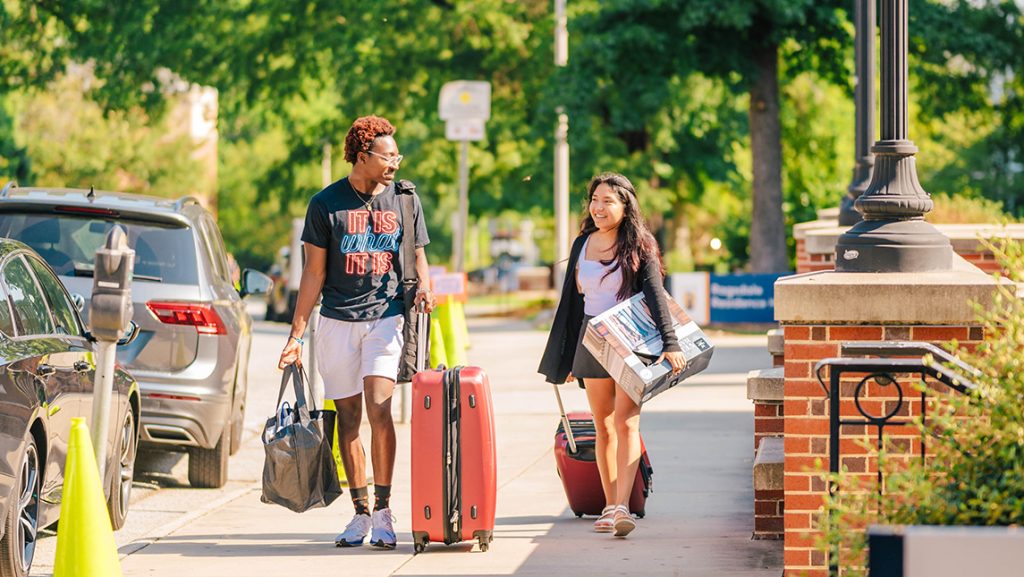 Two students walk down a sidewalk on campus carrying boxes and pulling suitcases.