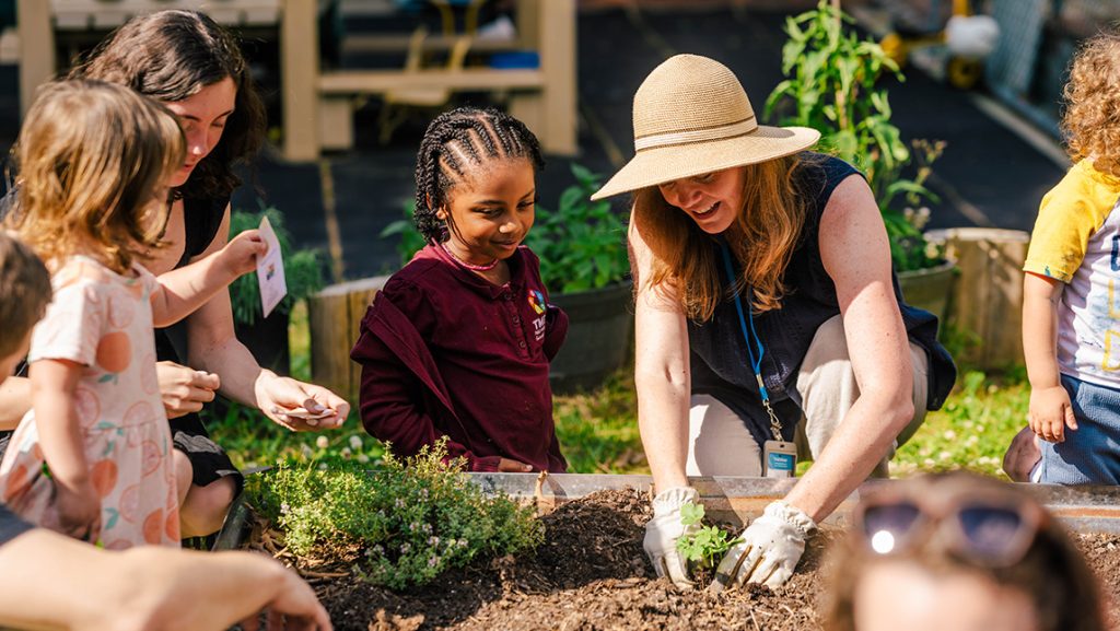 A Farm to ECE organizer shows a child how to plant vegetables in a UNCG garden.