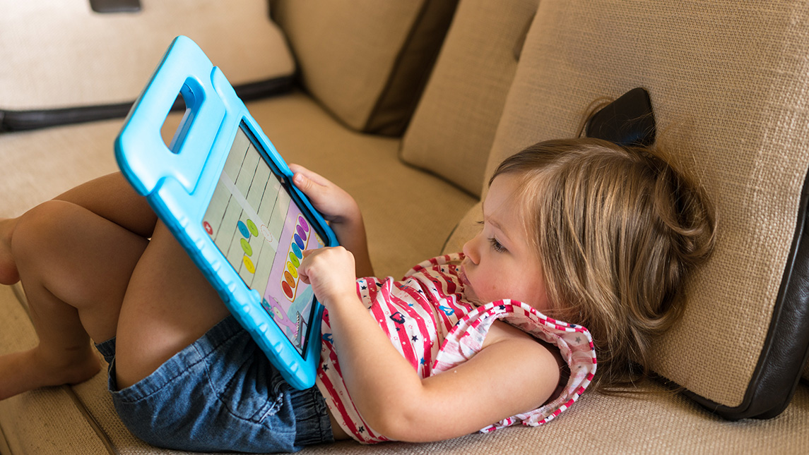 A toddler sits on the couch while playing games on a tablet.