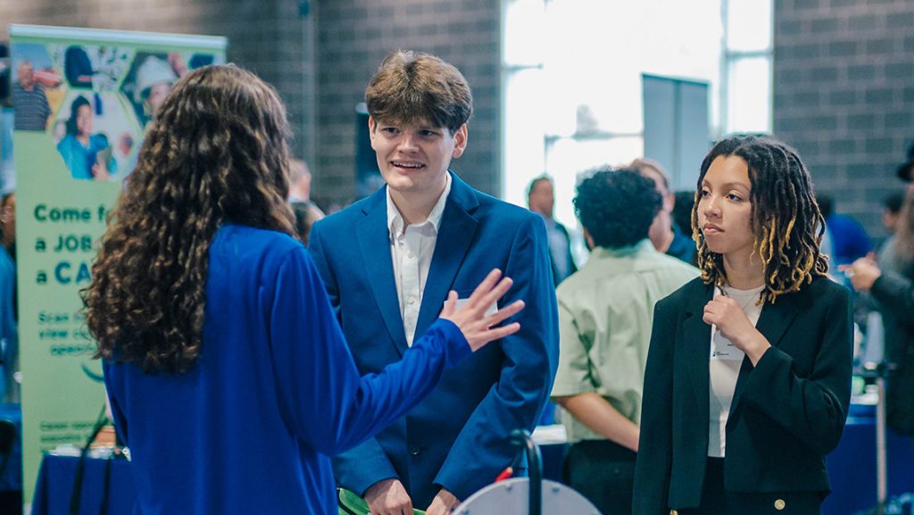 A male and female student in business suits chat with a rep at a career fair.