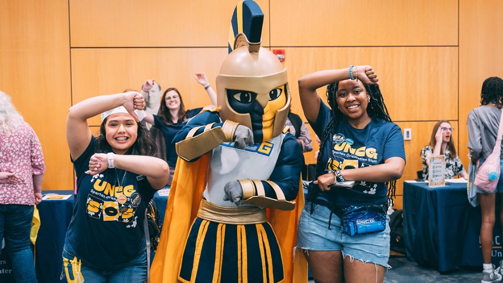 Two UNCG students make the "G" gesture with mascot Spiro.
