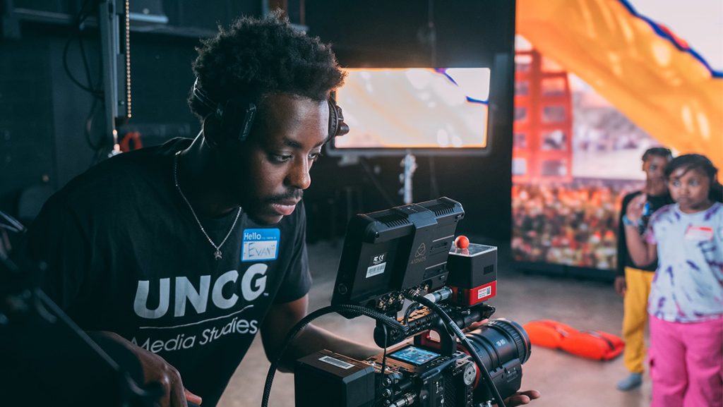 A UNCG media studies student looks through a camera at children who are performing.