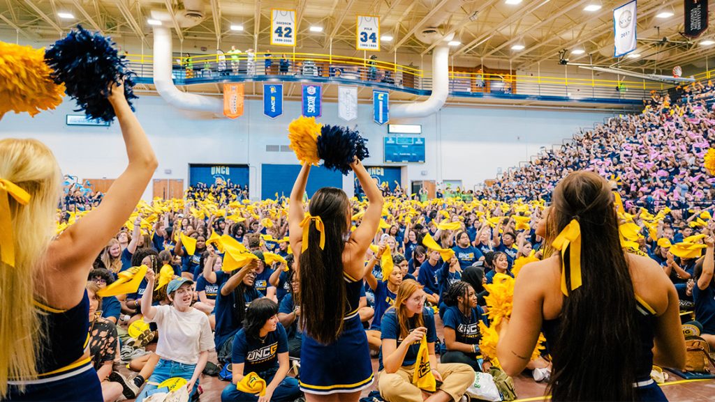 A gym full of students in UNCG shirts twirling rally towels as cheerleaders face them shaking pom-poms in the foreground.