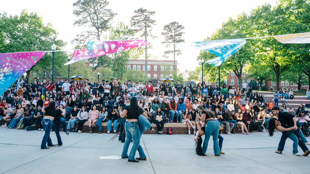 Students dance in pairs in front of Moran Commons with a crowd of students watching from the steps and campus buildings and trees in the background.
