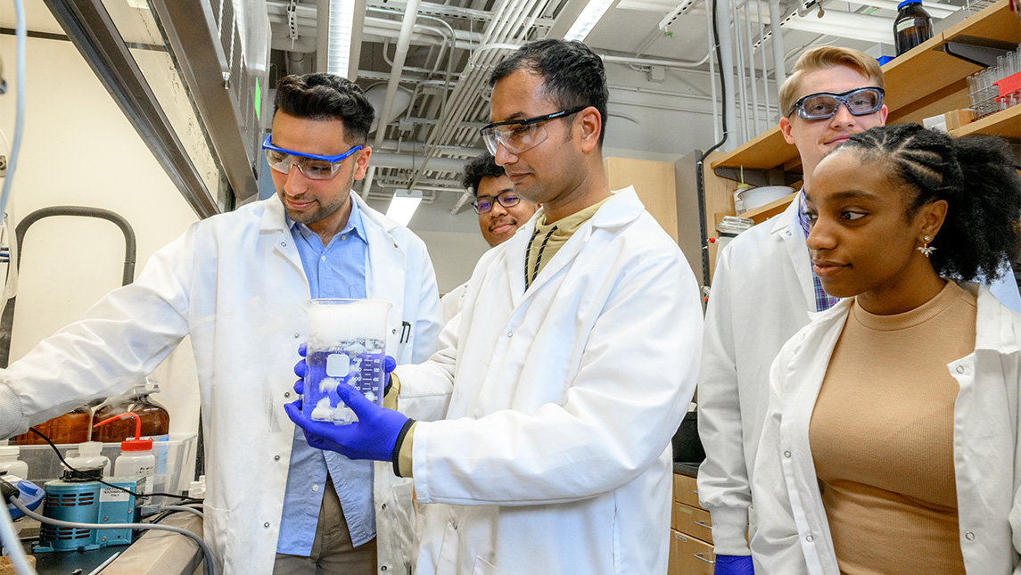 Students in UNCG Professor Mitch Croatt's lab gather around a sample in a beaker.
