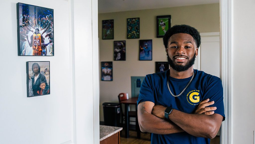 Student leans against door frame in a residence hall with framed sports photos on the wall behind him.