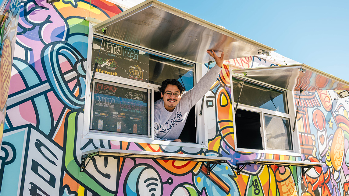 Man in UNCG sweatshirt leans out of a window of a colorful food truck to open it.