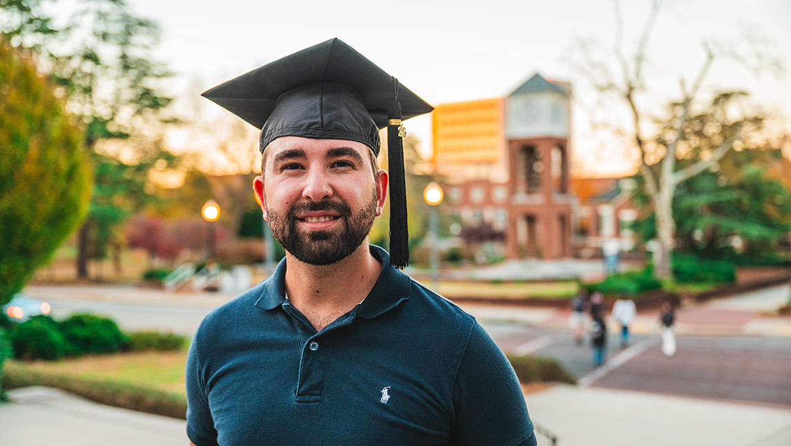 A man stands in a graduation cap on UNCG campus.