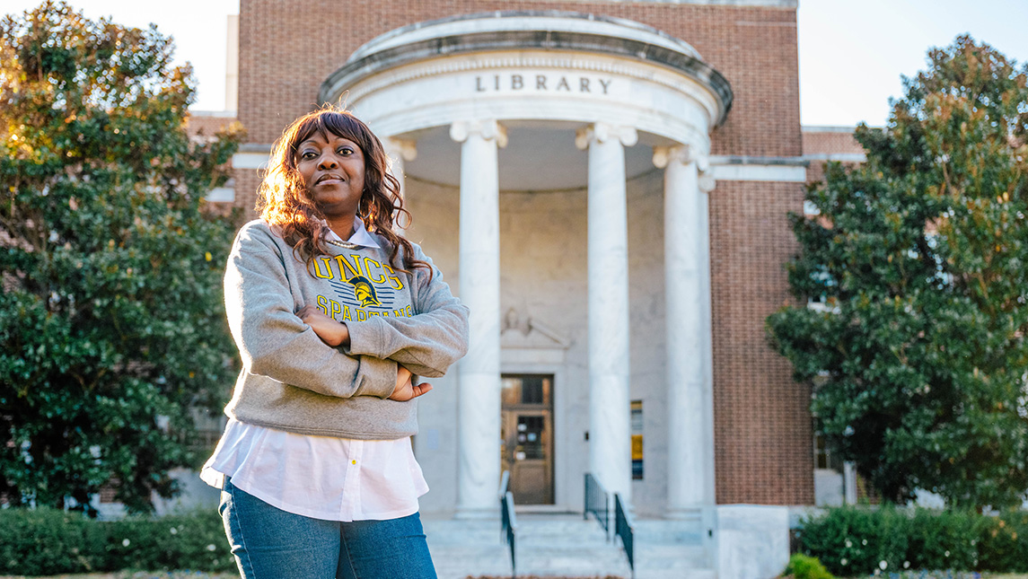 Woman in UNCG sweatshirt stands in front of the library.