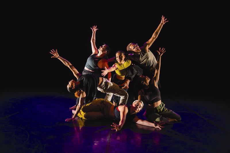 Dance troupe hits a final pose with outstretched hands on a darkened stage.