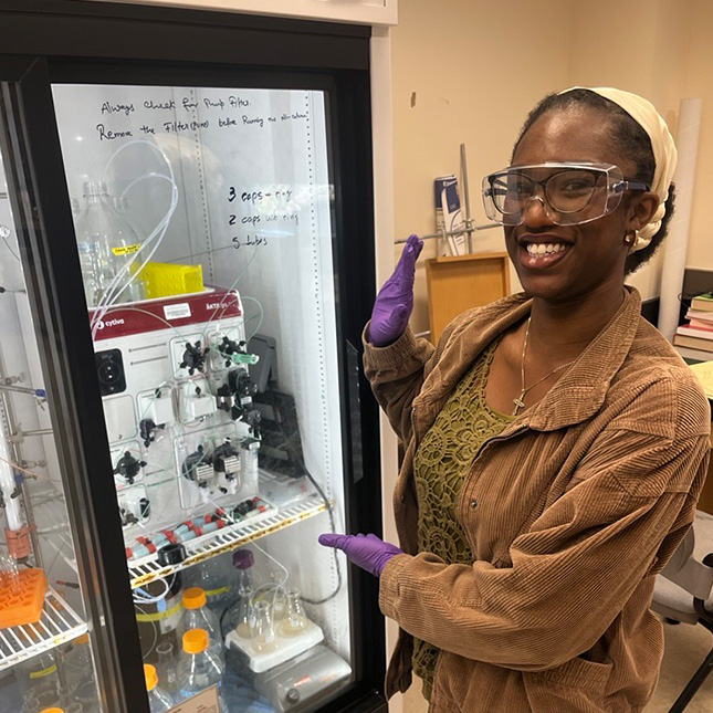 Student in safety googles stands near a frig holding lab samples.