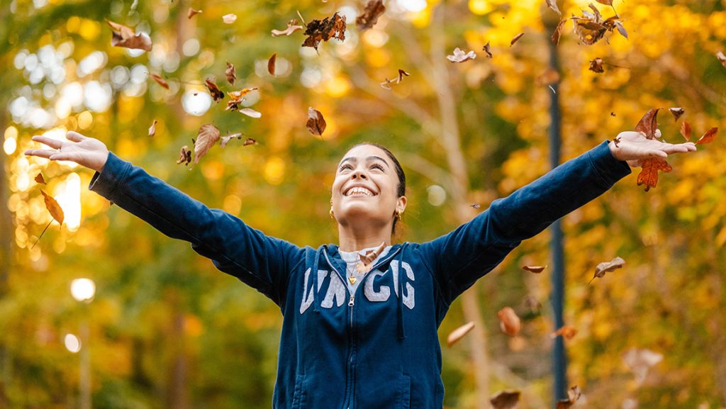 Student in UNCG sweatshirt with arms outstretched as she throws fall leaves in the air in the woods.