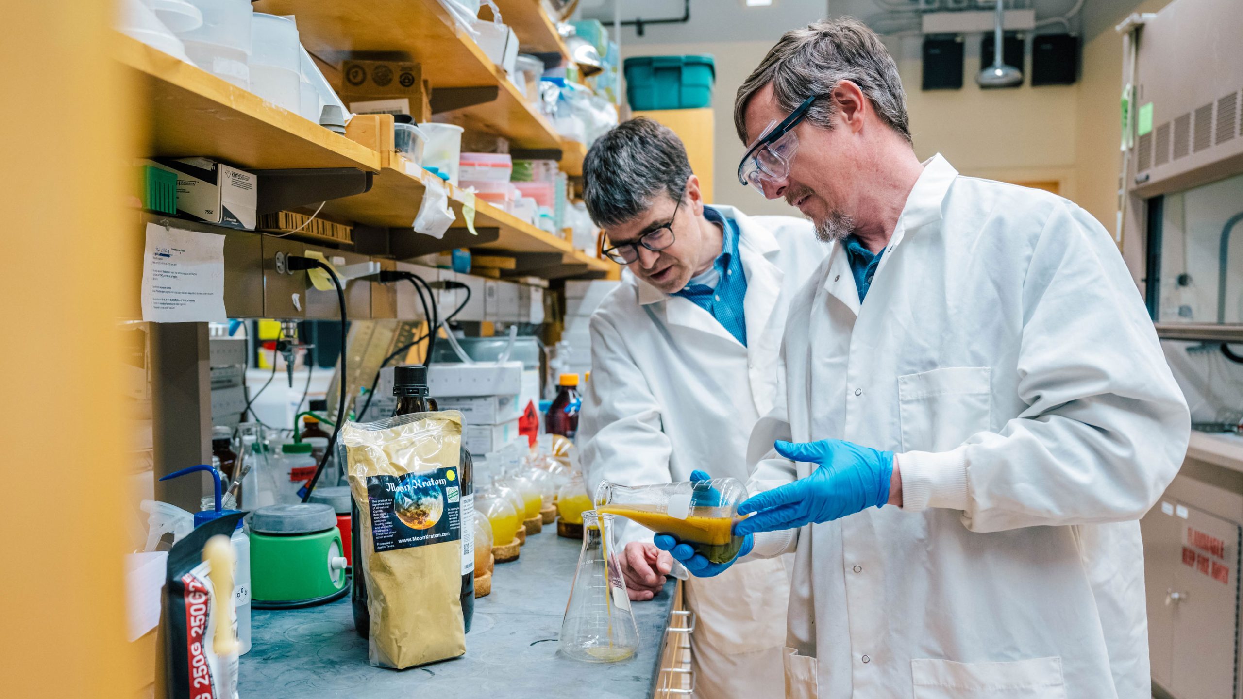 Two men in lab coats pour liquid into a flask in a lab.