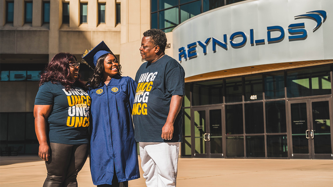 Graduate stands in cap and gown flanked by parents in UNCG t-shirts in front of the Reynolds American building.