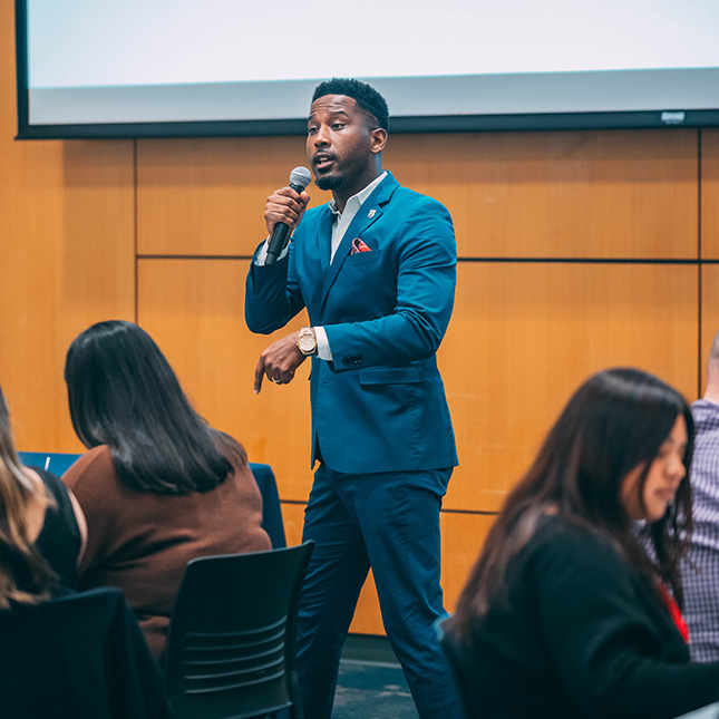 Speaker walks with a mic in front of students seated at tables. 