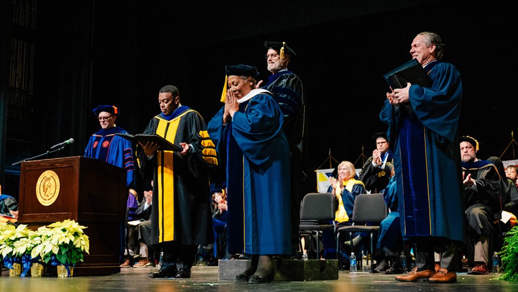 On a stage in the UNCG Auditorium, a woman is bestowed with Honorary Letters while other faculty members look on.