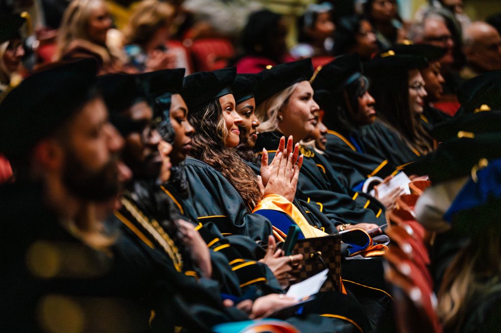 Doctoral candidates in cap and gown seated in an auditorium. One in the middle smiles and claps.