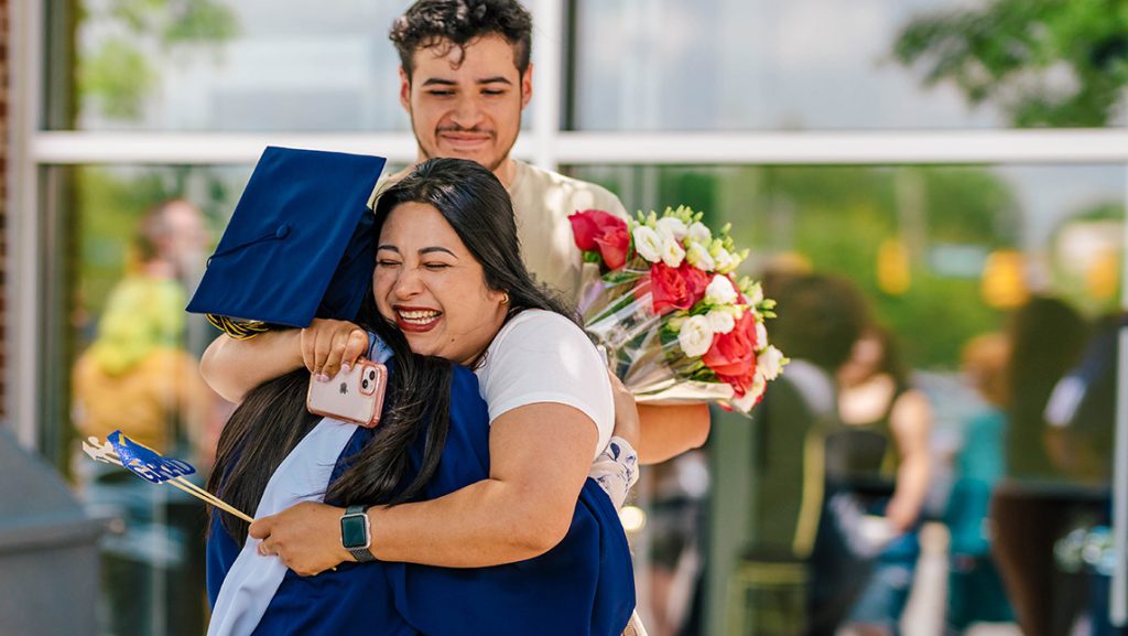 Parents with flowers hug their UNCG graduate in her cap and gown.