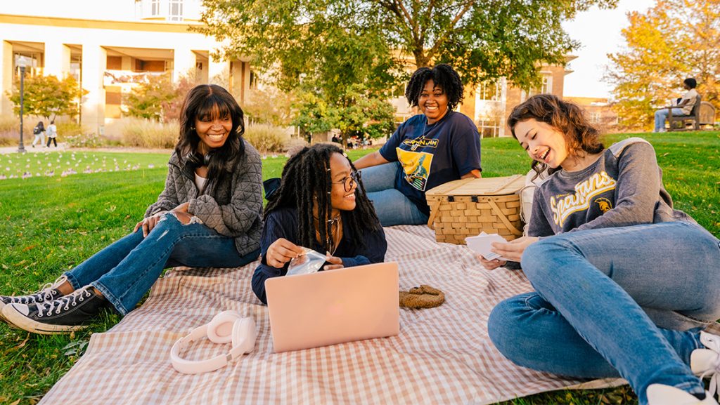 UNCG students sit on a blanket on the lawn around their laptop.