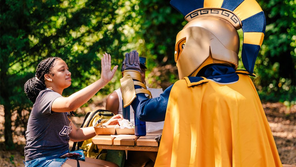 UNCG mascot Spiro gives a student at a picnic table a high five.