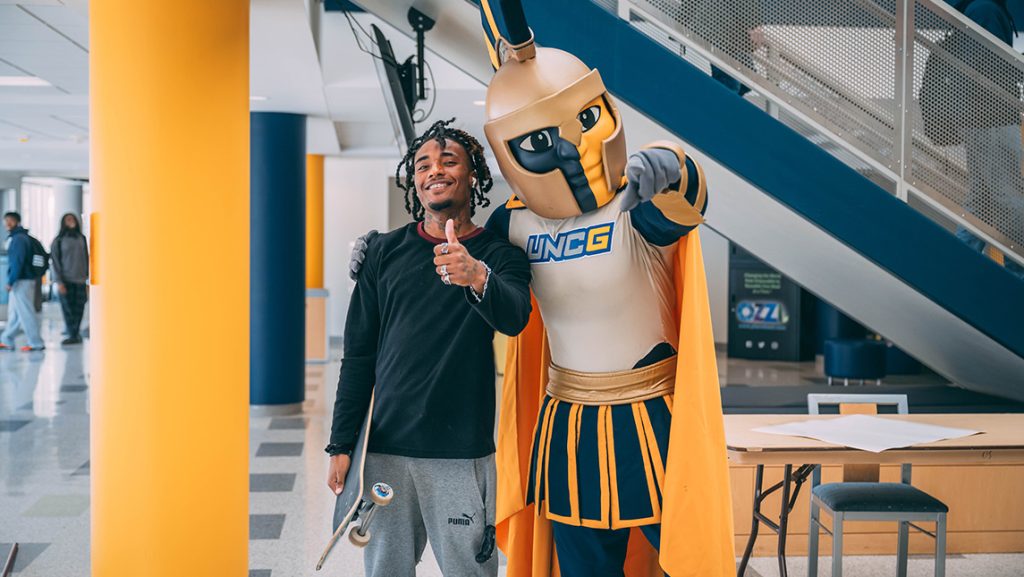 UNCG mascot Spiro poses with a student holding a skateboard.